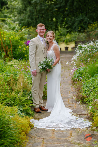 Newlyweds standing together on a flower-lined garden path at Askham Hall